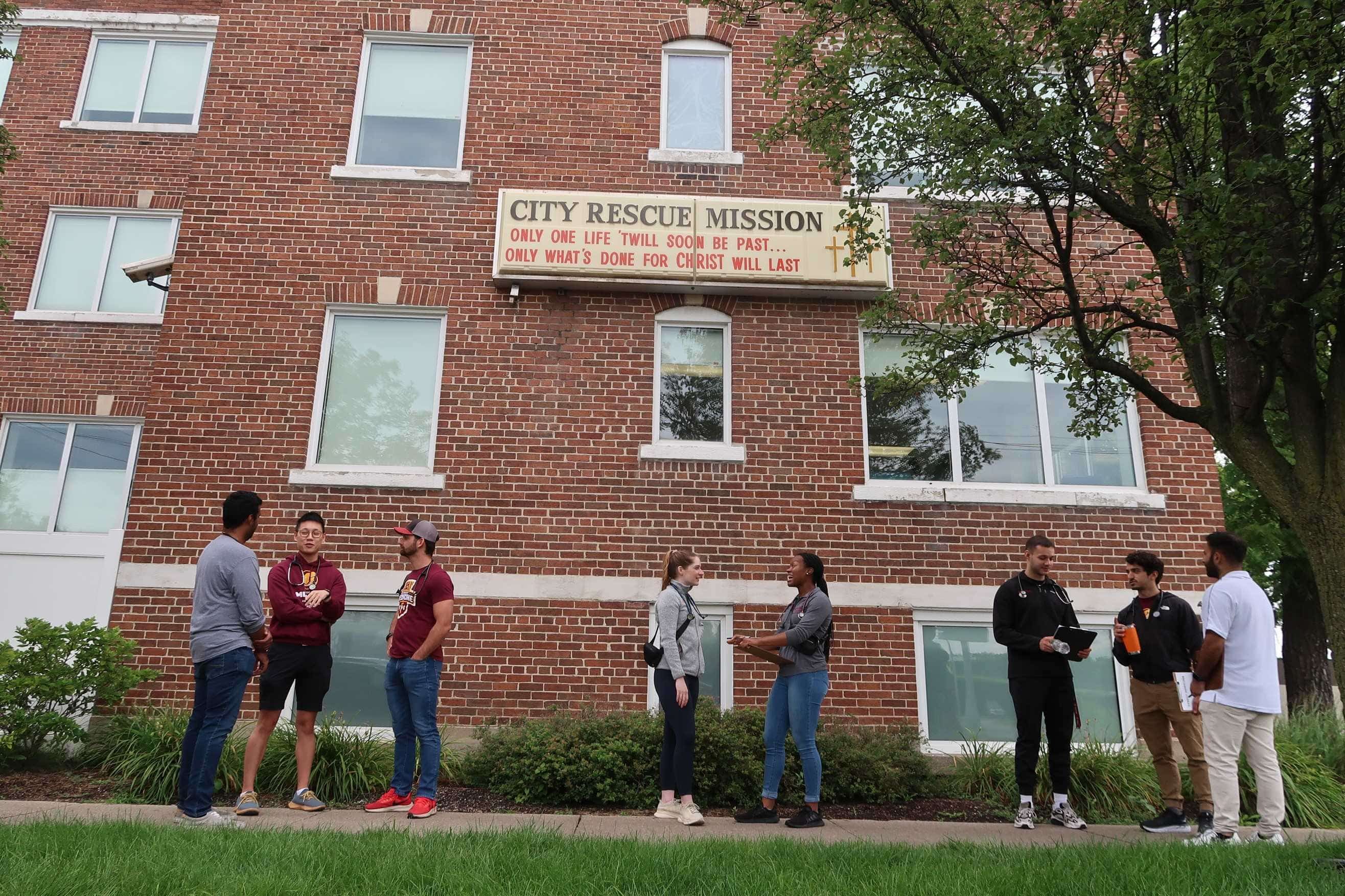 Eight college students dressed in casual clothing are talking with each other in front of a tall brick building.