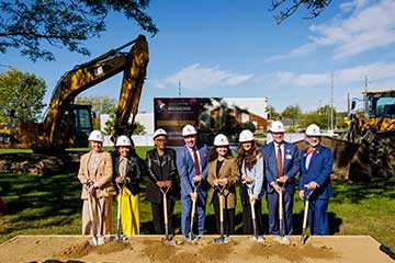 A group of 8 men and women in hard hats stand with shovels in the ground at a construction site.