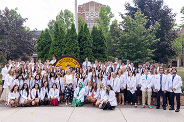 A large group of students in white coats gather around the University seal smiling at the camera.