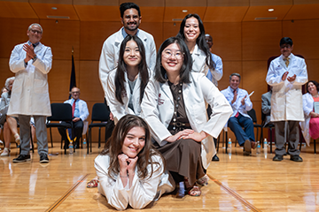 Medical students in white coats smile at the camera as medical school faulty applaud in the background.