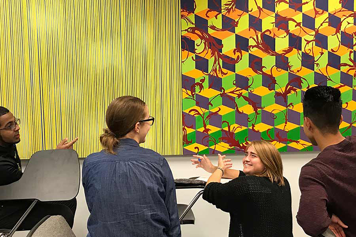Students in an Art History class at Central Michigan University gather in a classroom for a discussion with wall art in the background.