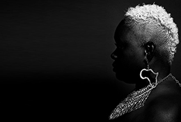 Black and white image of a black woman's profile. She has short cropped blonde hair, an earring shaped like the continent of Africa and a large ornate necklace