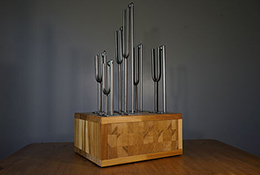 A sculpture. The base is a wooden box. Attached to the top of the box are various sized tuning forks.