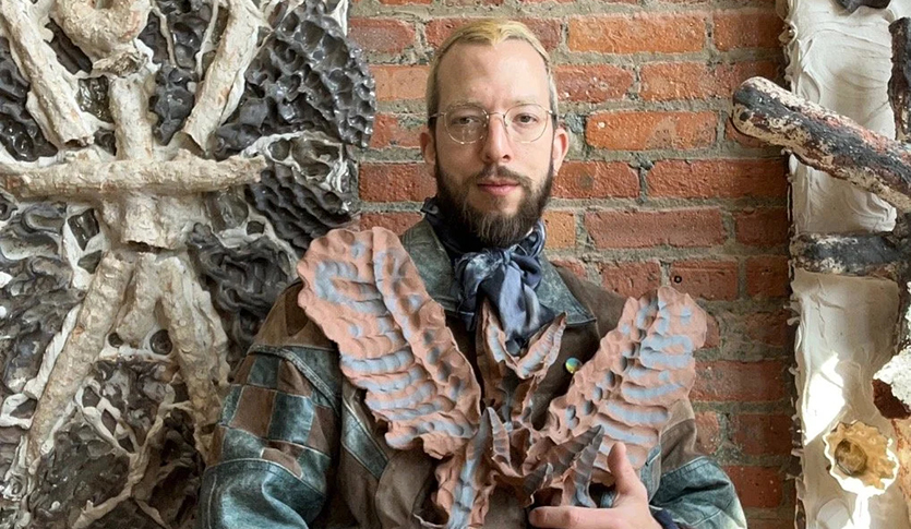 Dove Hornbuckle standing in front of a brick wall, holding one of their sculptures. There are sculptures hanging on the wall behind them as well.