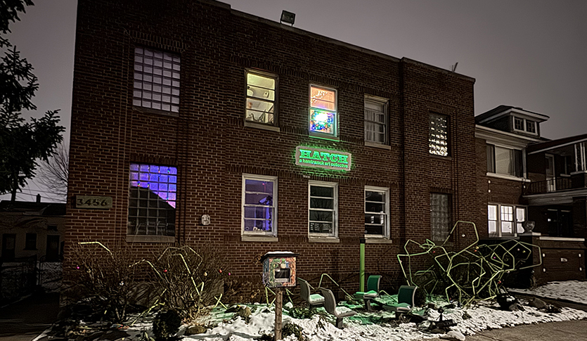 Exterior of a brick building. It has a couple lights seen through it's second floor windows. There's a neon sign glowing on the front of the building.