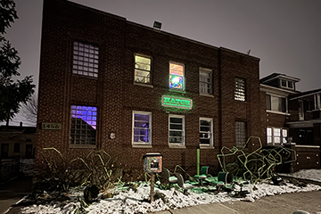 Exterior of a brick building. It has a couple lights seen through it's second floor windows. There's a neon sign glowing on the front of the building.