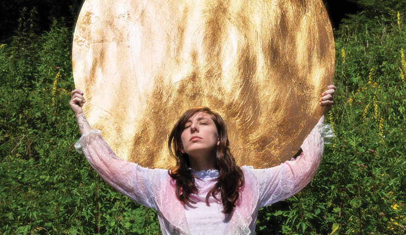 Artist Danielle Owensby is holding a large golden disc up behind and above her head. The sun is reflecting off of it. There is a wall of greeenery behind her.