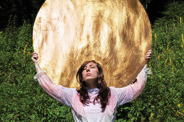 Artist Danielle Owensby is holding a large golden disc up behind and above her head. The sun is reflecting off of it. There is a wall of greeenery behind her.