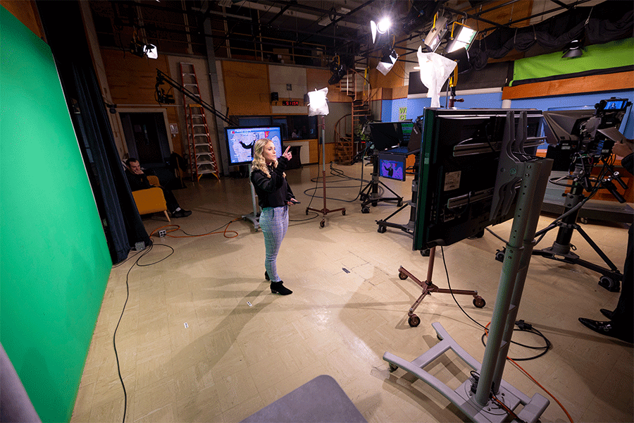A broadcast student stands in the studio in front of a green screen, as she practices giving a weather update with screens all around her and cameras capturing her.