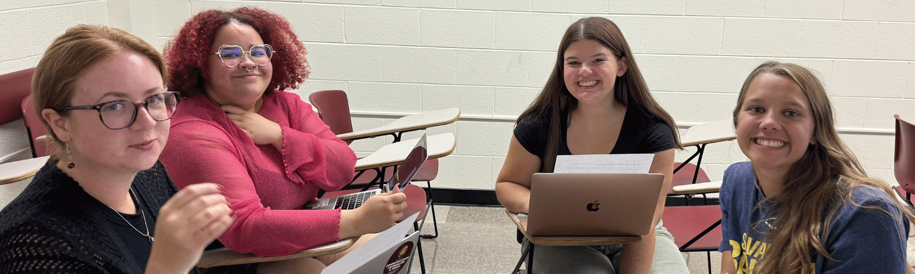Four students sitting in a classroom, sitting at desks while smiling and laptops are on top of each desk.
