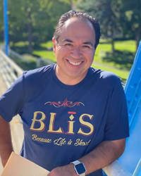 Headshot of John Gonzalez wearing a blue t-shirt with green trees in the background.