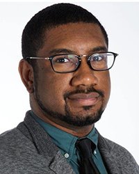 Headshot of Khristopher Brooks wearing a dark green shirt and black tie in front of a white backdrop.