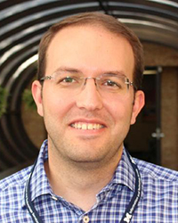 Headshot of Kyle Cozad wearing a plaid shirt in front of a door on a brick wall.