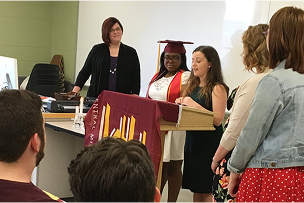 Department of Communication students giving a group speech in front of a classroom with a Central Michigan University flag on a podium in the College of Art and Media.