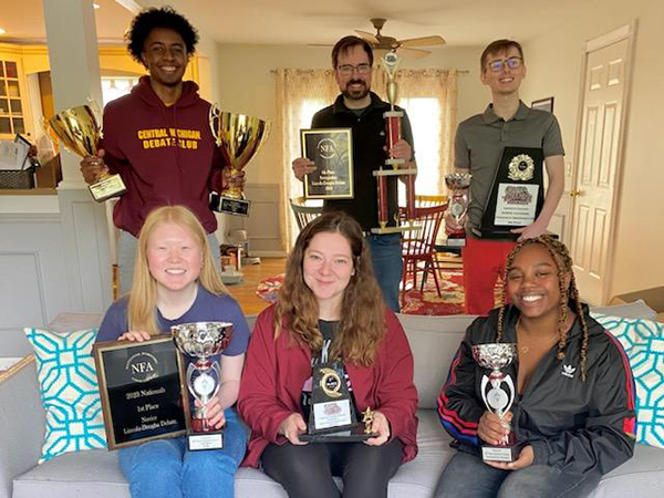 Six students are posing with the many trophies they won at the last debate tournament.