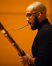 Headshot of Carlos Clark on a golden backdrop. He is playing his bassoon.