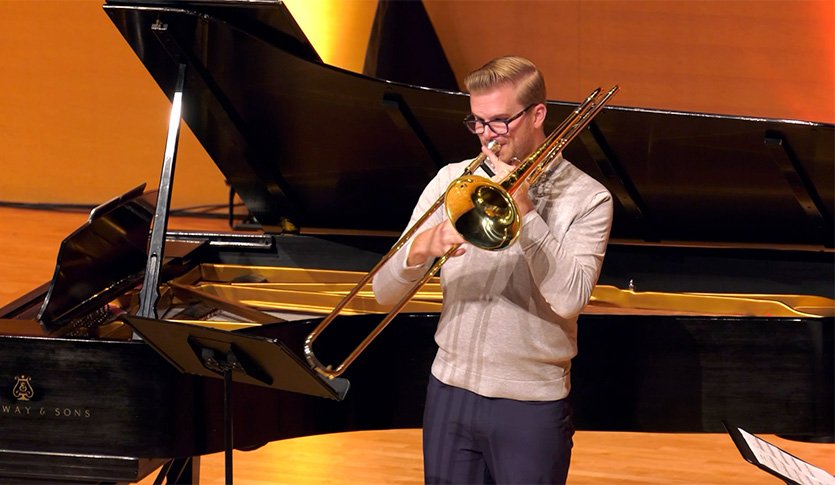 A picture of Dr. Cole Bartels wearing a beige sweater and playing trombone at Staples Family Concert Hall.