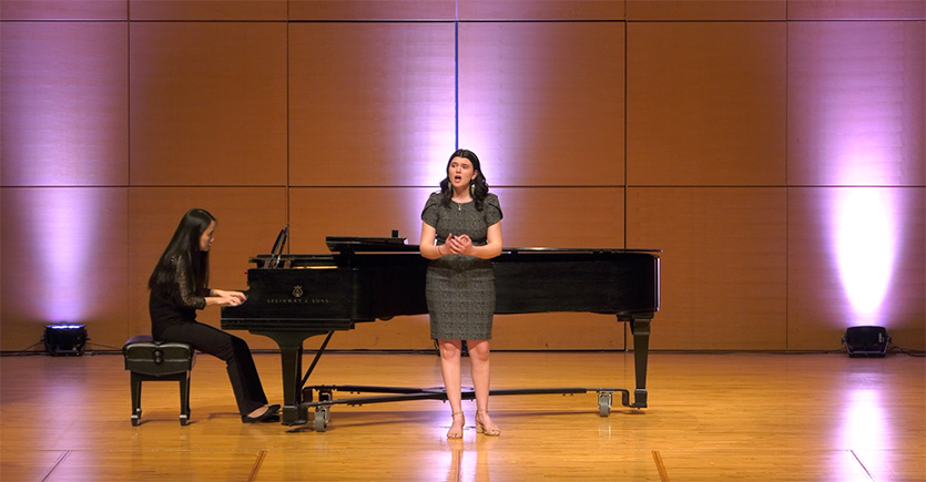 A student wearing a dark gray dress sings in front of a grand piano on stage at Staples Family Concert Hall.