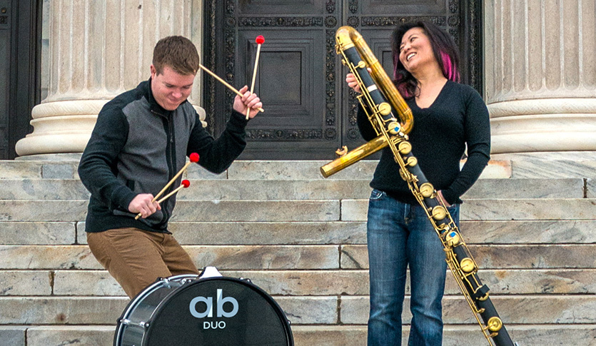 A man playing a drum and woman playing bass flute posing on a staircase.