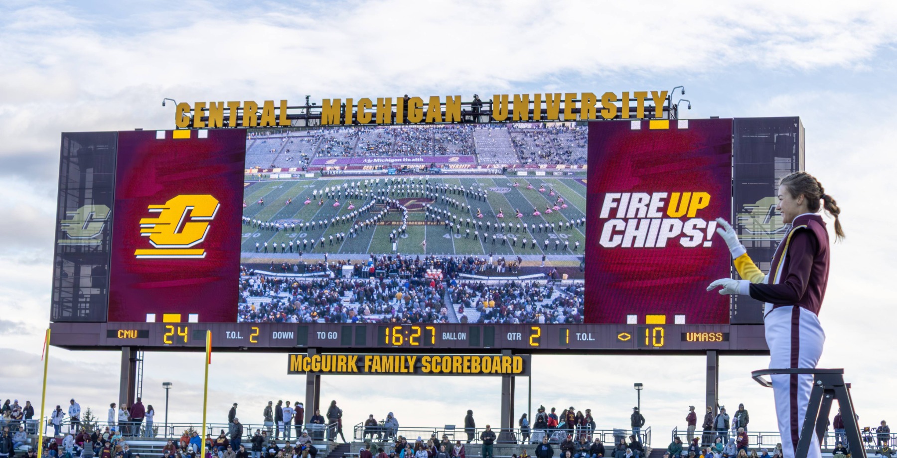 a large scoreboard with a football field and people watching with the Chippewa Marching Band drum major in front