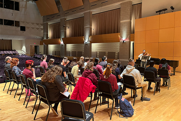A picture of students sitting on stage with another student playing viola on the background