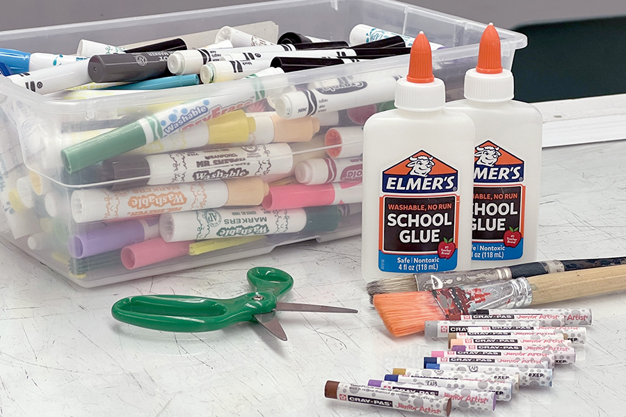 A desk with a box of markers, scissors, bottles of glue, paintbrushes and oil crayons on it.