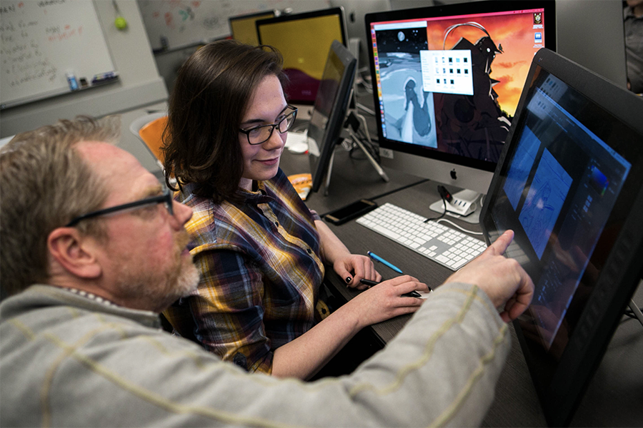 a student sits at a computer while a professor points at the screen