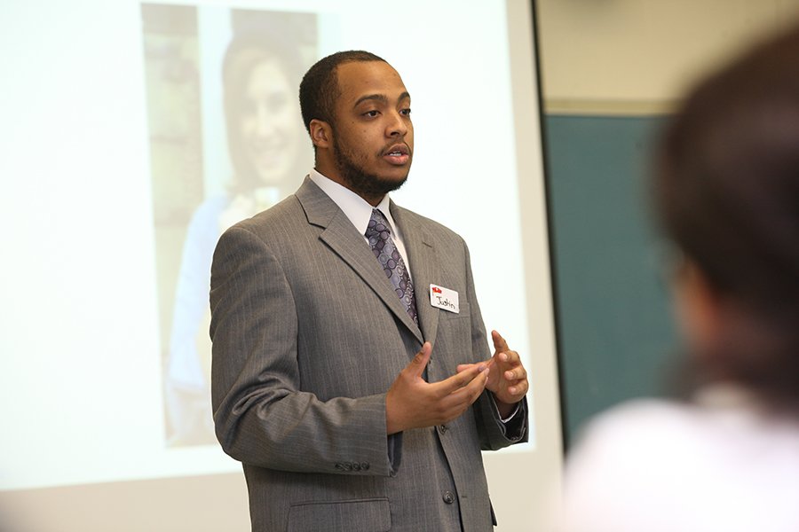 A student stands at the front of the room, in front of a projector screen, giving a presentation.