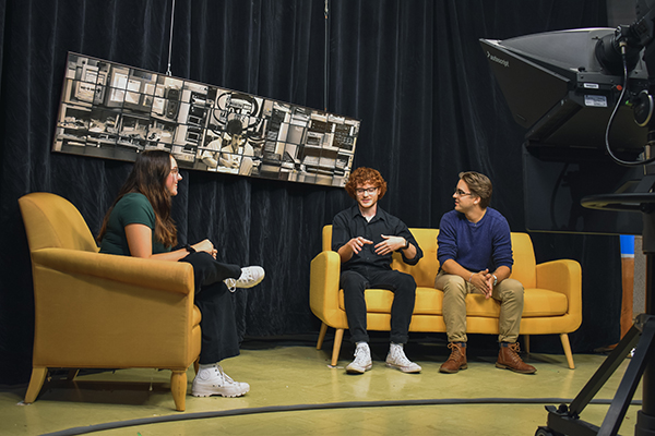 One student interviews two other students on a set with couches.