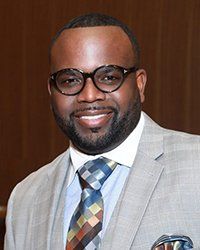 Headshot of Lester Booker with a brown pin striped wall behind him.