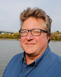 Headshot of Tom Henry. You can see a body of water behind him with trees along the horizon.