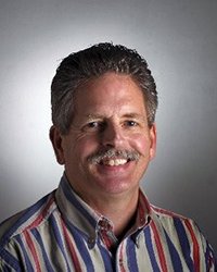 Headshot of Steve Morse wearing a red, grey and blue collared shirt against a soft grey backdrop.