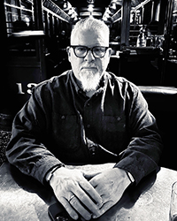 20250626_MHoF_PSgriccia_001_200x250 Greyscale headshot of Phil Sgriccia. He's sitting at a table in a restaurant with his hands folded on the table.