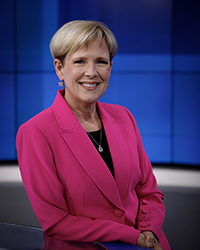 Headshot of Ann Emmerich standing at a news desk in a bright pink blazer with a blue backdrop.