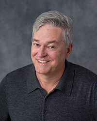 Headshot of Andrew Heller wearing a dark grey shirt in front of a soft grey backdrop.