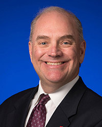 Headshot of Tim Pollard smiling in a black suit and maroon tie in front of a bright blue backdrop.