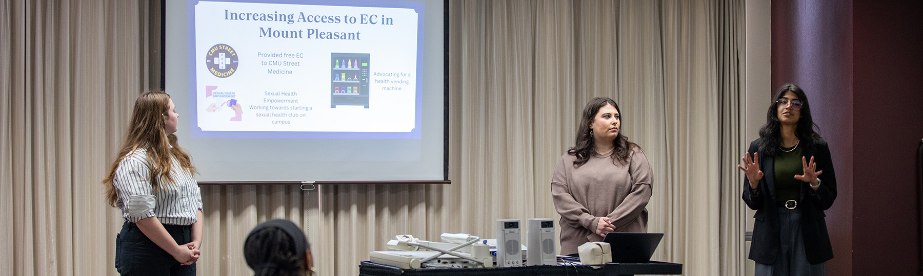 Three women dressed professionally stand in front of a projector while giving a presentation on access to EC in Mount Pleasant.