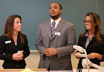 Three students stand at the front of a classroom, presenting to the room.