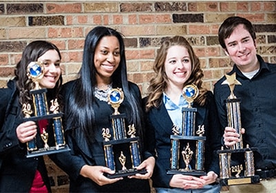 Four students pose in front of a brick wall with the trophies they won at a debate competition.