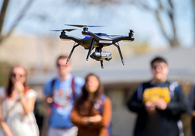 A flying drone in the foreground while a group of students watch in the background.