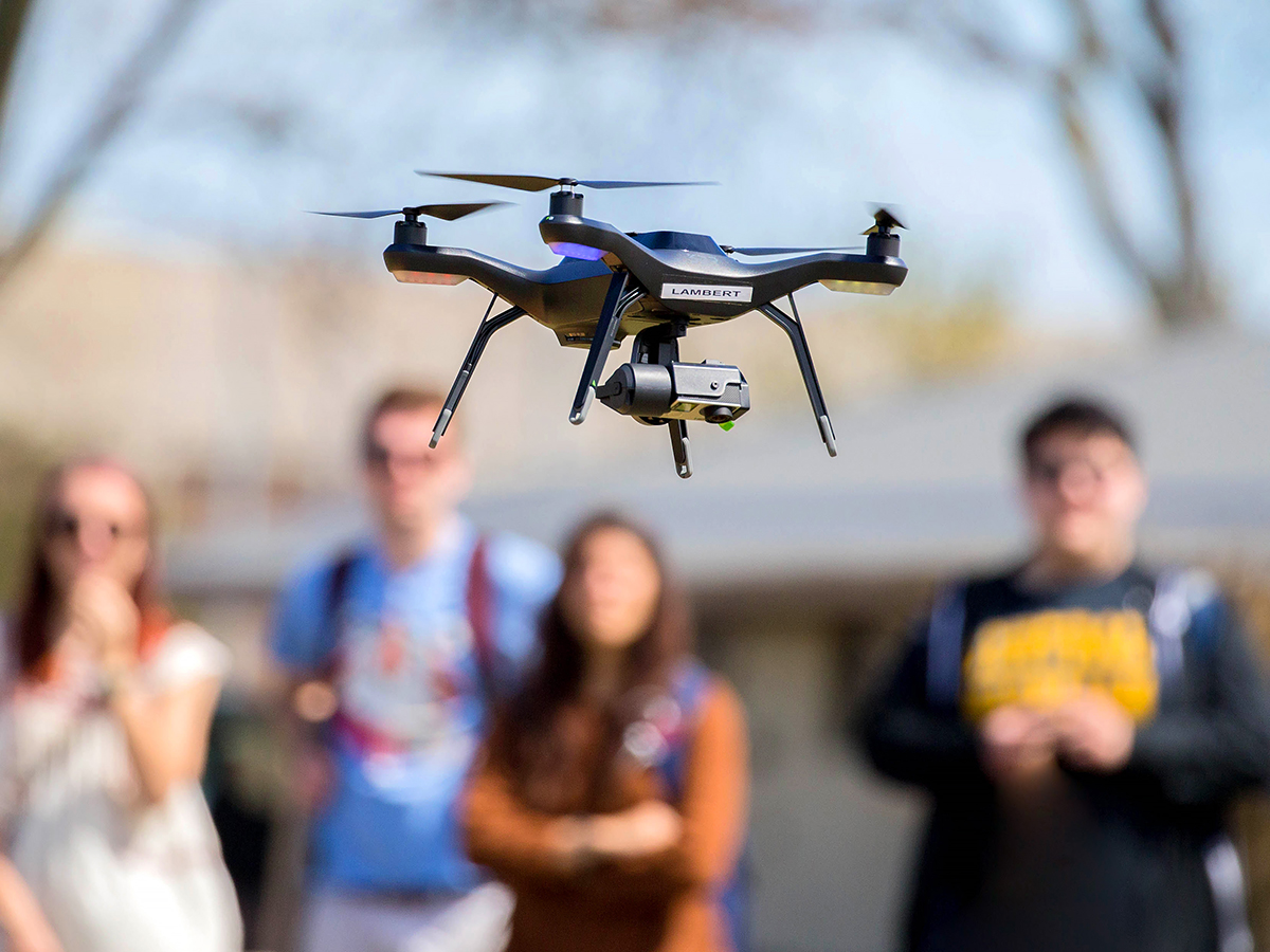 A flying drone in the foreground while a group of students watch in the background.