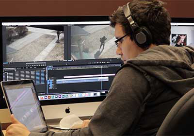 A student works on his film, sitting at a desk with several monitors and editing software.