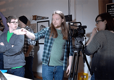 Students in the middle of a film production. One student is behind the camera, another is explaining the scene, while a third is listening to the conversation.