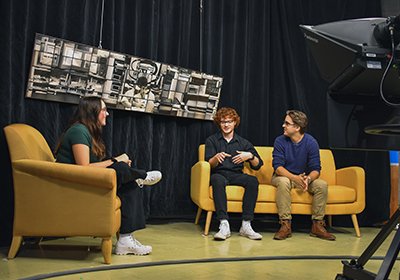 Students are interviewing each other for a television show. They are sitting on couches on a stage.