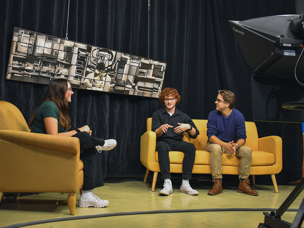 Students are interviewing each other for a television show. They are sitting on couches on a stage.