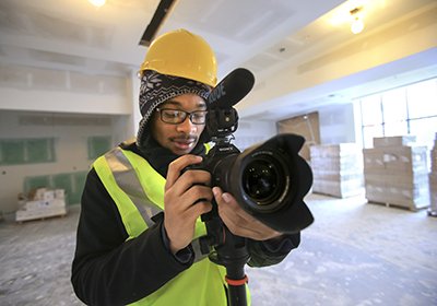 Photojournalism student taking pictures in a construction area. He is wearing a reflective vest and hard hat.