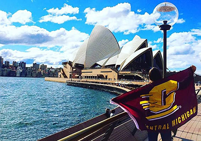 A student stands on a bridge while wearing a CMU flag as a cape. The Sidney opera house in Australia is in the background.