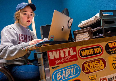 A student is working at a computer at a desk in the radio station on campus.
