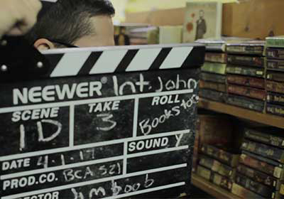 Close up of a movie slate. An actor in front of a shelf of books can be seen behind the slate.