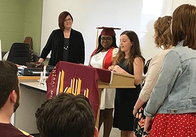 Students and staff are standing at the front of a classroom presenting a scholarship to a fellow student.